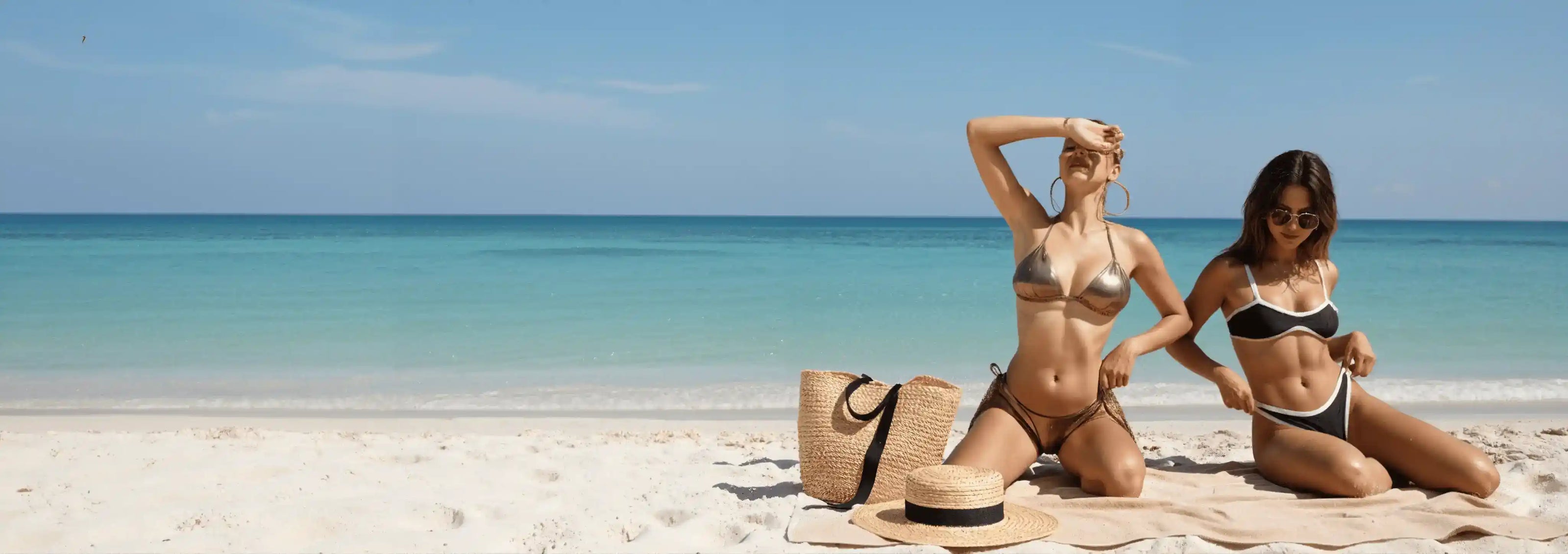 Two women in bikinis sitting on a beach with clear blue water and sky.